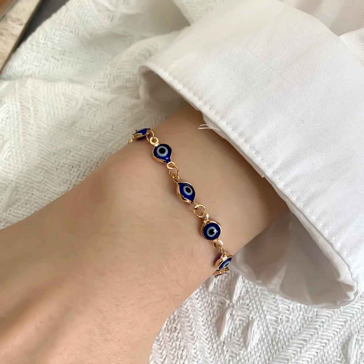 A close-up of a gold charm bracelet featuring blue evil eye charms worn on a wrist against a white fabric background.