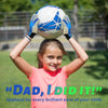 A smiling girl holds up a soccer ball with blue and white goalie gloves above her head in an outdoor setting.