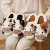 A pair of white cartoon cow winter home slippers with brown and black accents are shown being worn by a person.