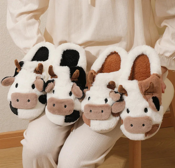 Une paire de pantoufles d'hiver en forme de vache blanches, avec des accents marron et noir, portées par une personne.