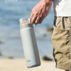 A hand holds a light gray insulated stainless steel water bottle outdoors near the sea.