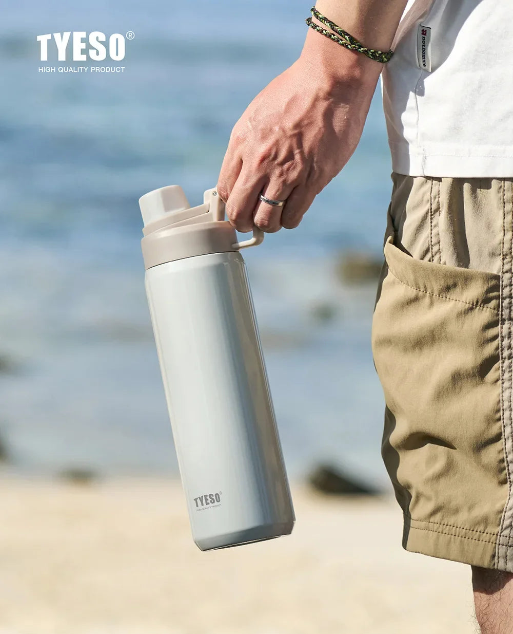 A hand holds a light gray insulated stainless steel water bottle outdoors near the sea.