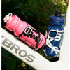 Una botella de agua para ciclismo duradera rosa y azul, apoyada sobre una superficie blanca al aire libre.