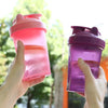 A hand holds a pink leak-proof sports shaker bottle against a blurred green outdoor background.