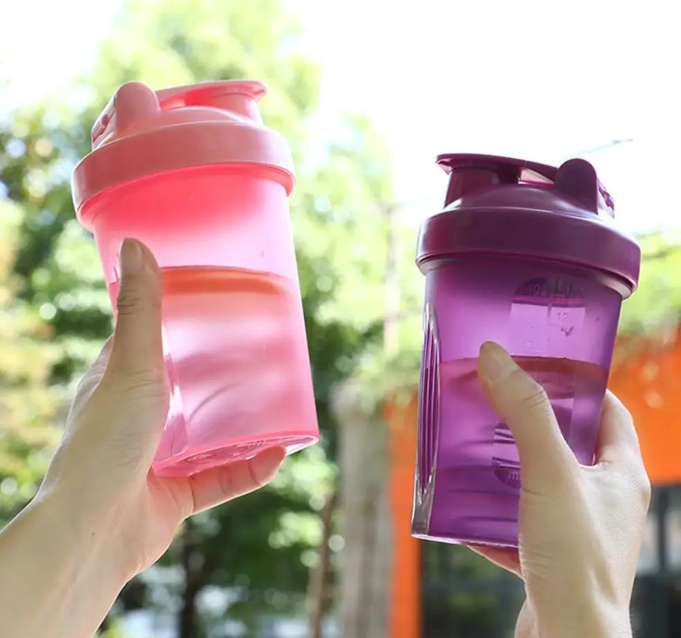 A hand holds a pink leak-proof sports shaker bottle against a blurred green outdoor background.
