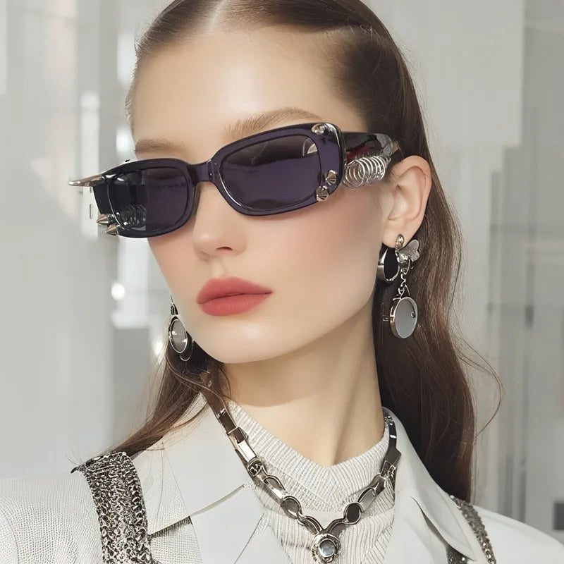 A close-up of a woman wearing dark purple spiked rivet sunglasses and a silver necklace against a light background.