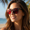 A woman wearing dark red sunglasses outdoors with palm trees in the background.