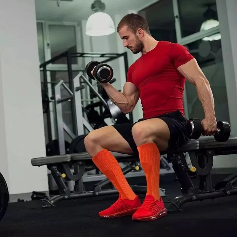 رجل مفتول العضلات يرتدي قميصًا أحمر وشورتًا أسود يؤدي تمرين رفع أثقال مع جوارب ضاغطة برتقالية زاهية