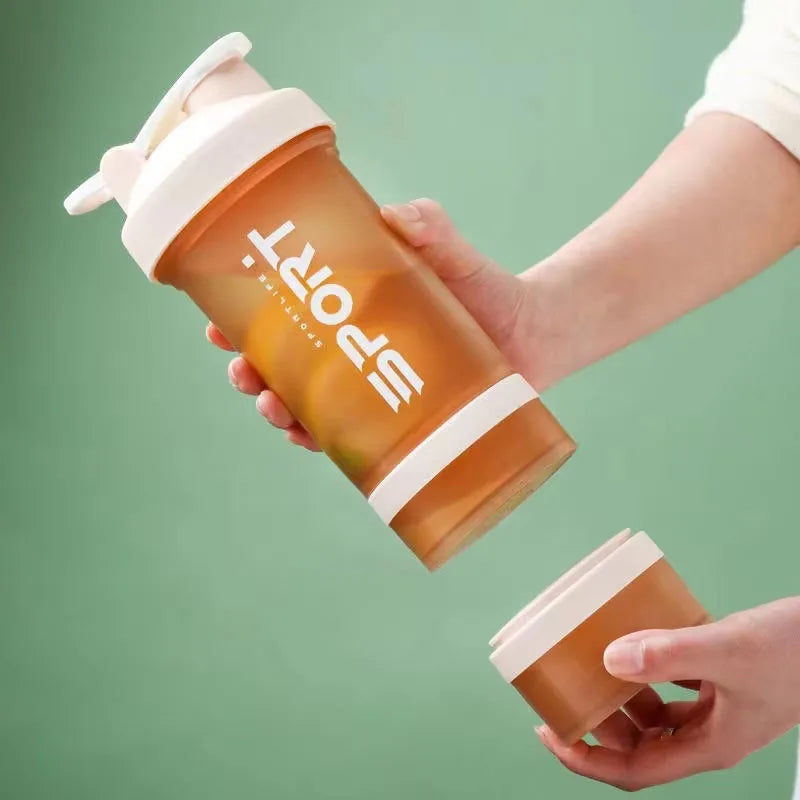 A hand is holding an orange-colored sports shaker bottle with a pink lid against a green background.