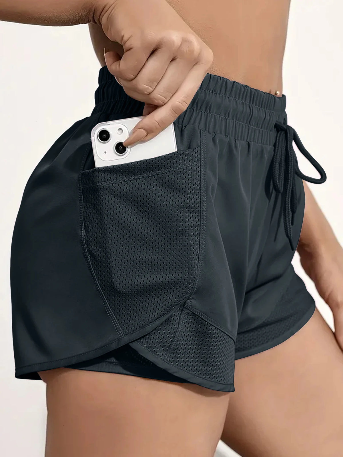 A close-up of dark gray athletic shorts with mesh pockets, showing a hand placing a smartphone into a pocket.