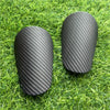 Pair of black carbon fiber-look soccer shin guards resting on green grass.