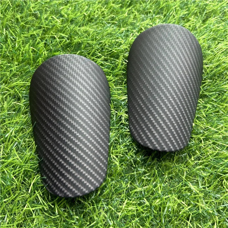 Pair of black carbon fiber-look soccer shin guards resting on green grass.