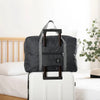 A black foldable travel duffel bag with a carrying strap is placed on a white suitcase next to a bed.