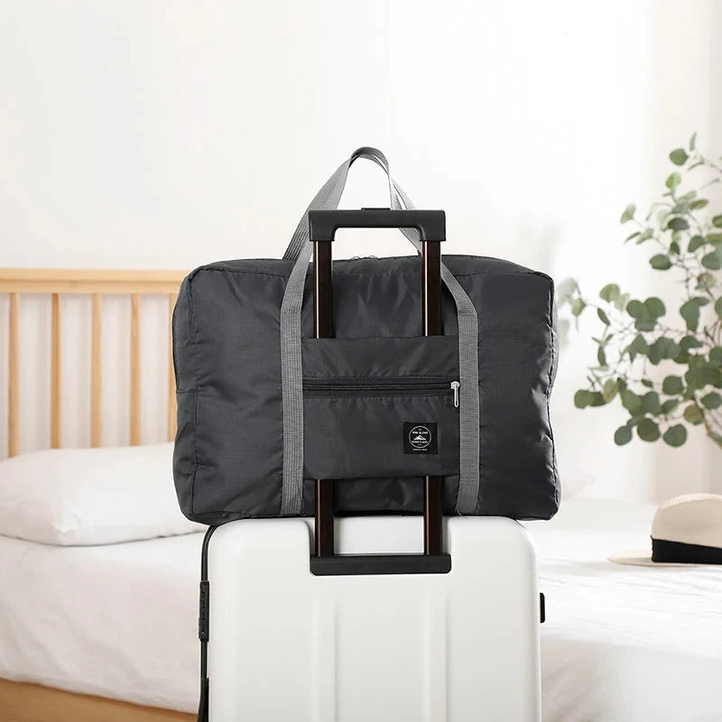 A black foldable travel duffel bag with a carrying strap is placed on a white suitcase next to a bed.