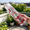A hand holds a clear reusable water bottle with a strawberry design, featuring the text Sweet Cherry, outdoors.