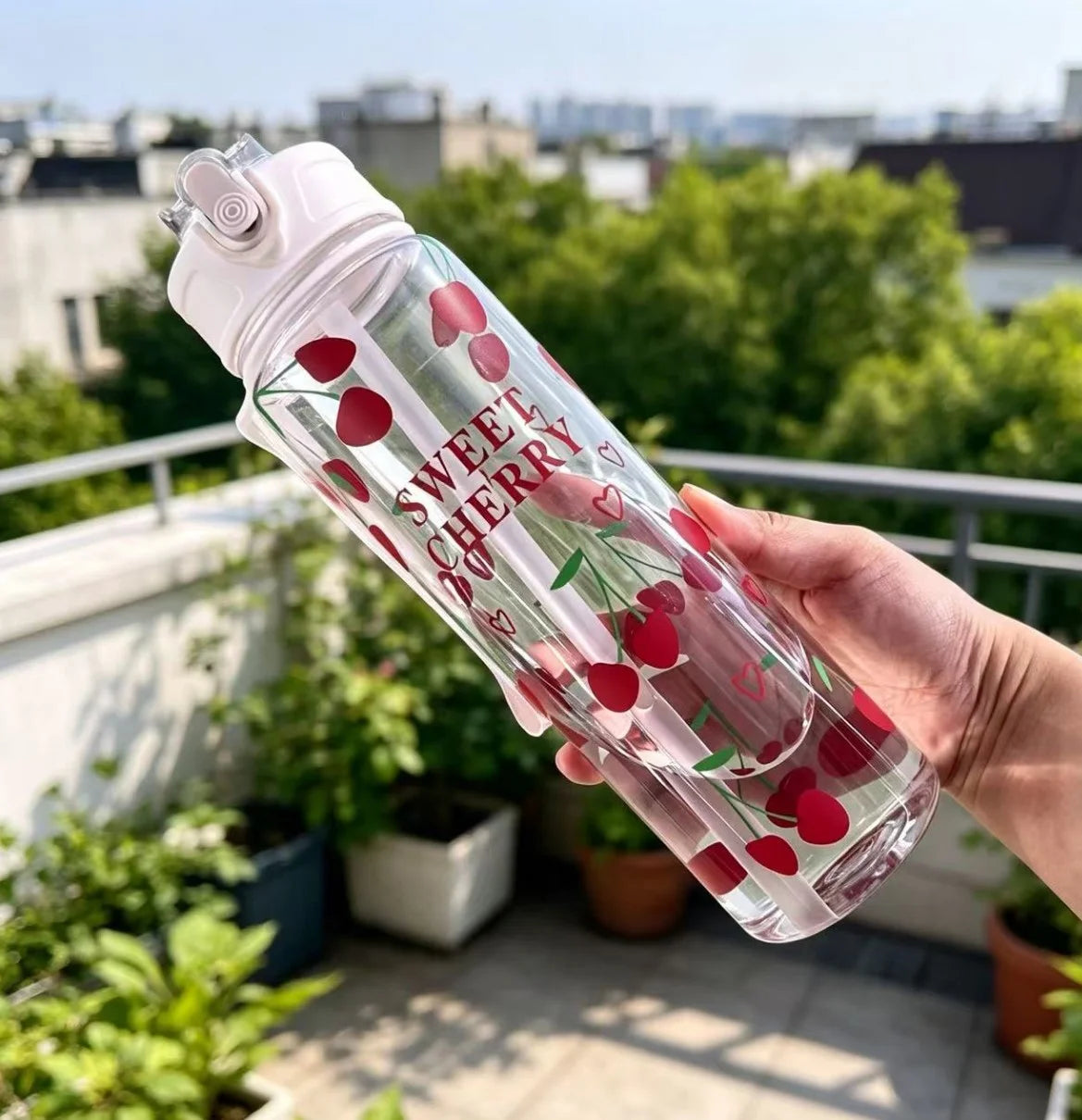 A hand holds a clear reusable water bottle with a strawberry design, featuring the text Sweet Cherry, outdoors.