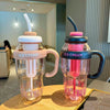 Two clear reusable water bottles with straws and handles, colored pink and dark gray, sitting on a wooden surface.