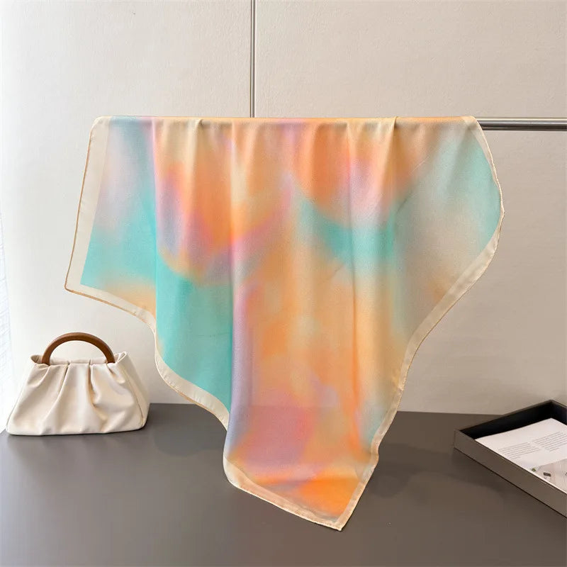 A sheer silk scarf featuring a soft watercolor blend of peach, orange, and teal colors draped on a gray surface.