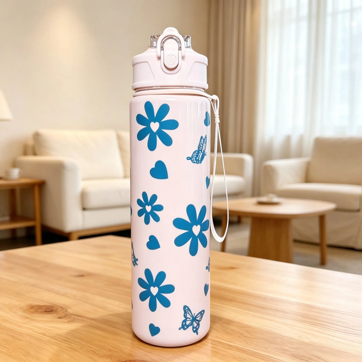 A pink reusable water bottle with blue floral and butterfly designs sits on a wooden table in a bright living room.