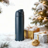A dark blue insulated stainless steel tumbler with a straw lid sits in the snow next to wrapped gifts and a decorated Christm