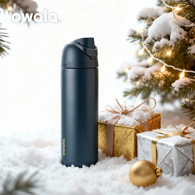 A dark blue insulated stainless steel tumbler with a straw lid sits in the snow next to wrapped gifts and a decorated Christm