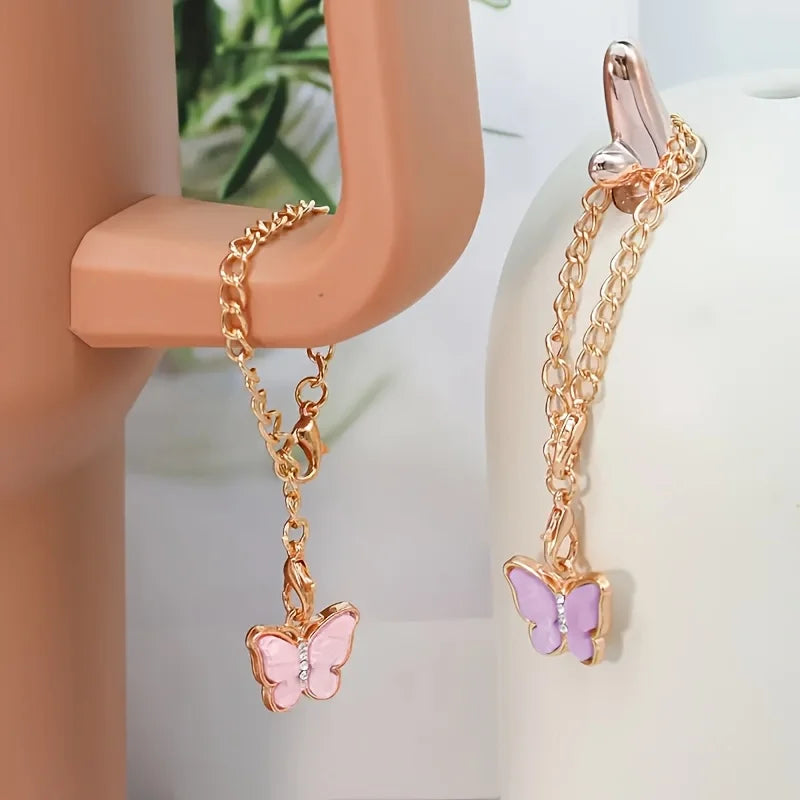 A gold-colored butterfly charm necklace is displayed on a pink holder, next to a similar piece with a rose gold butterfly cha