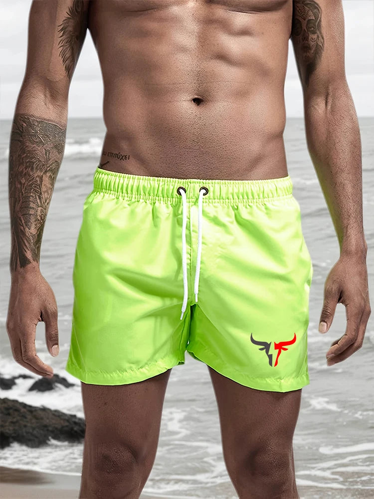 A muscular man wearing neon yellow quick-drying swim shorts standing near the ocean.
