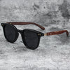 Black sunglasses with dark lenses and a brown wood grain frame resting on a gray textured surface.