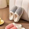 A pair of cozy plush fleece slippers in white and gray are resting on a wooden floor next to a cream-colored sofa.