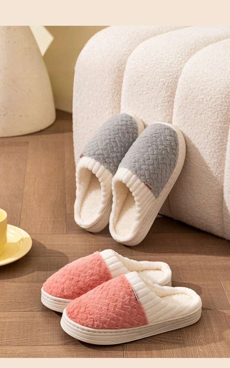 A pair of cozy plush fleece slippers in white and gray are resting on a wooden floor next to a cream-colored sofa.