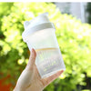 A hand holds a clear, leak-proof shaker bottle with a white cap against a bright green, leafy background.
