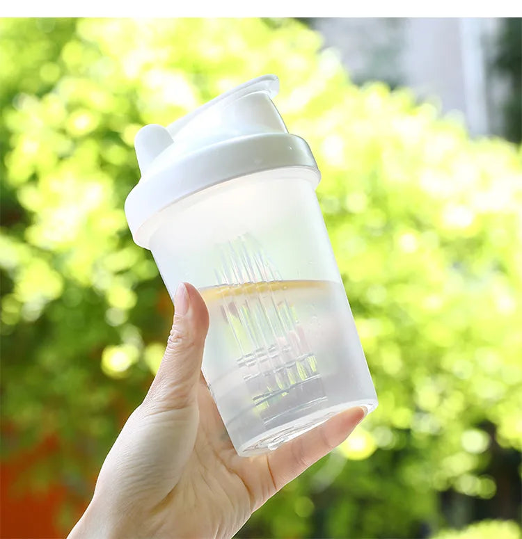 A hand holds a clear, leak-proof shaker bottle with a white cap against a bright green, leafy background.