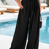 A woman models black flowy high waist drawstring wide leg pants outdoors near a swimming pool.