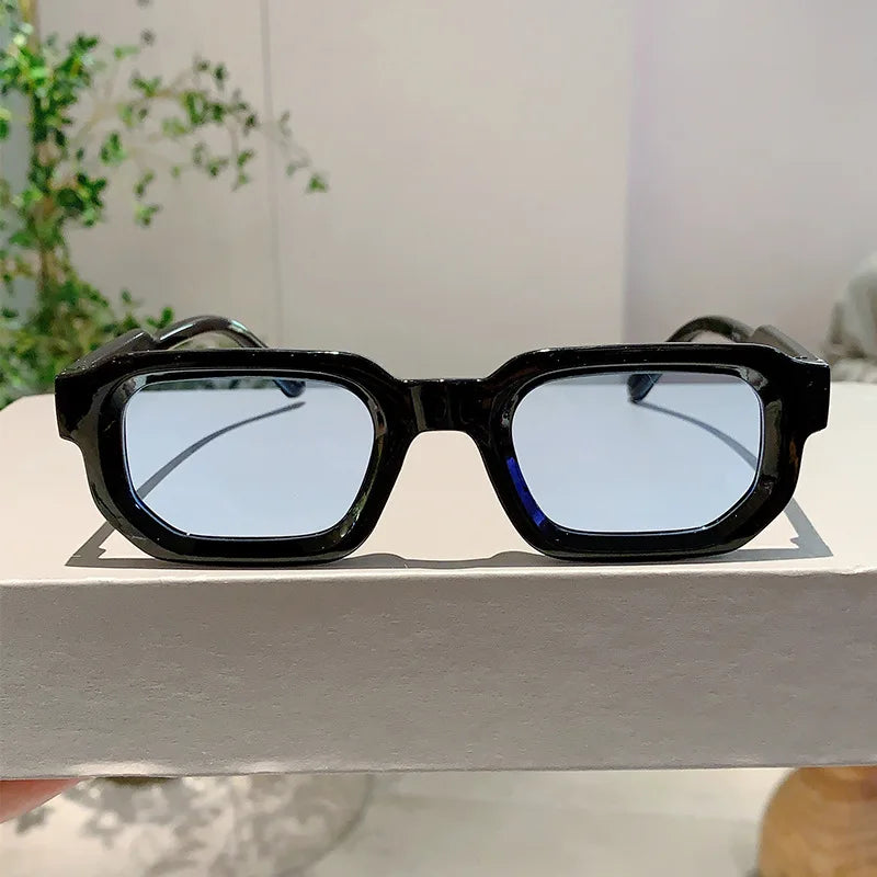 Lunettes de soleil à monture rectangulaire noire rétro posées sur une surface blanche.