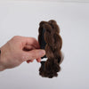 A hand is holding a dark brown twisted braided wig headband against a white background.