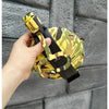 A hand holds a yellow and green patterned silicone camping kettle against a gray brick wall.