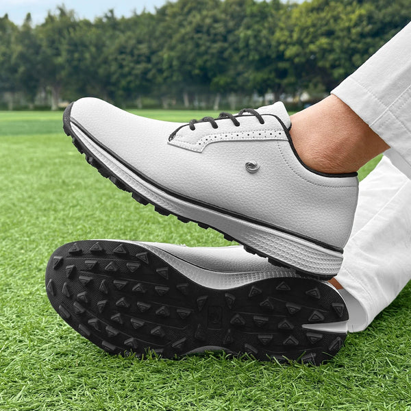 A close-up of a pair of light gray men's golf training shoes with black outsoles resting on green grass.