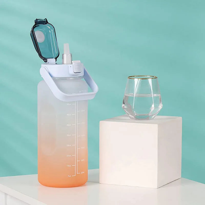Una botella de agua aislada motivacional de 2L transparente con tapa de pajita junto a un vaso sobre una superficie blanca.