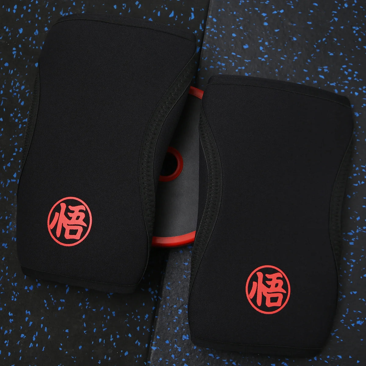 Une paire de genouillères de levage noires avec un logo rouge sont présentées sur une surface sombre.