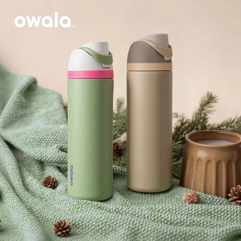 Eine hellgrüne isolierte Wasserflasche mit rosa Deckel und beige Flasche wird auf grünem Stoff neben Tannenzweigen präsentier