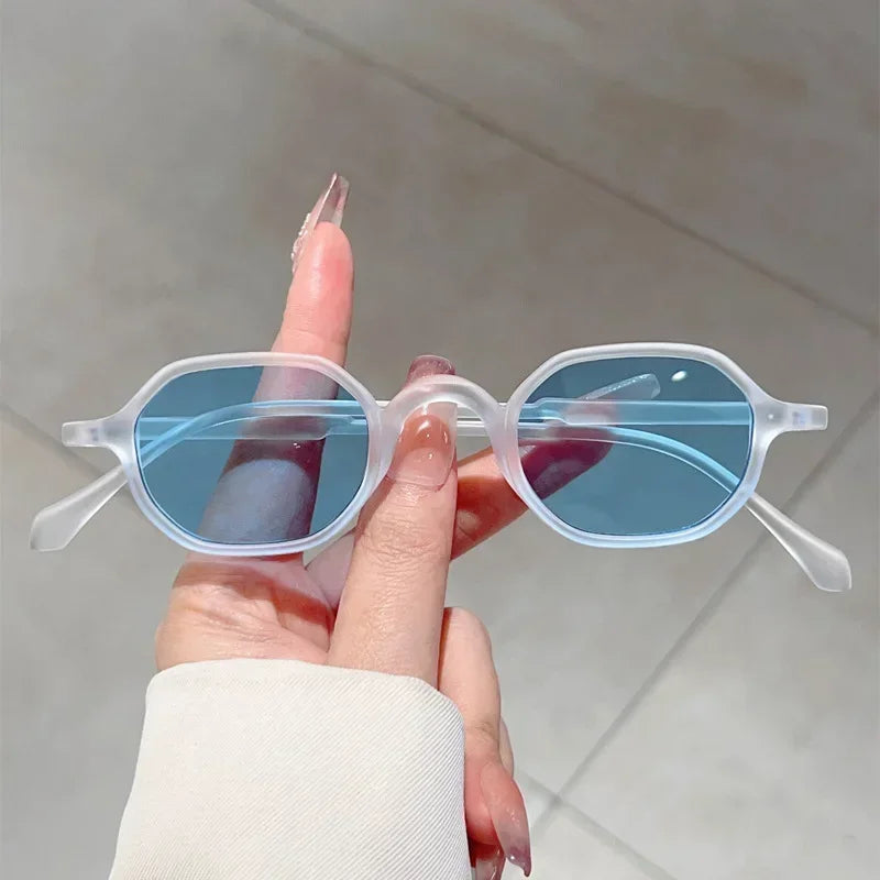 A hand holds a pair of clear-framed blue-tinted oval sunglasses against a light background.