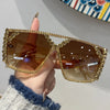 A hand holds a pair of gold-framed geometric rhinestone fashion sunglasses with brown lenses.
