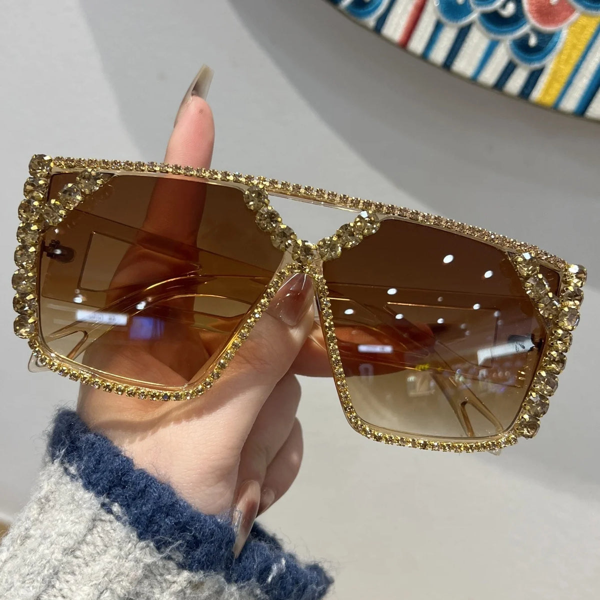A hand holds a pair of gold-framed geometric rhinestone fashion sunglasses with brown lenses.
