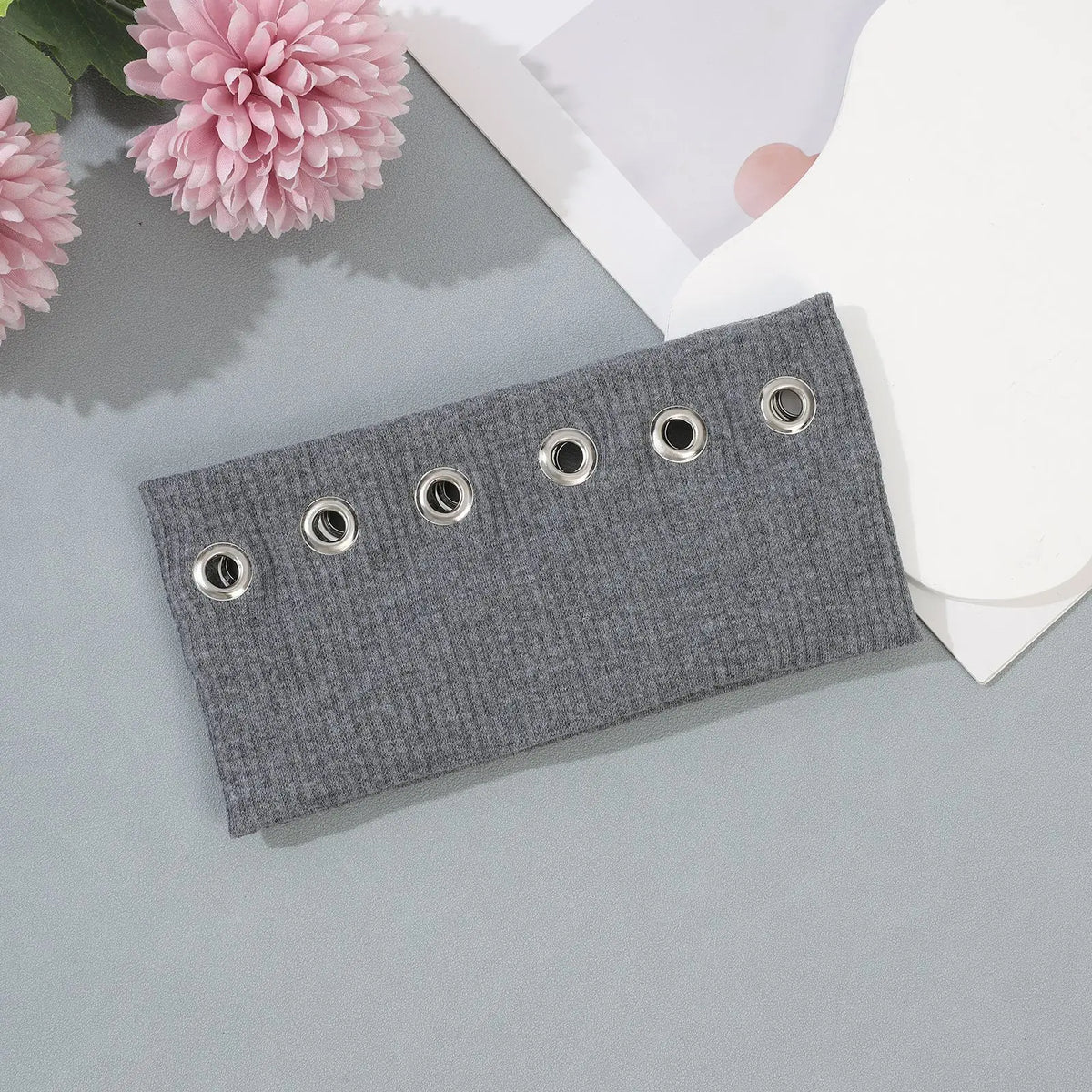 Un bandeau à œillets gris avec des grommets argentés, exposé sur une surface gris clair à côté de fleurs roses.
