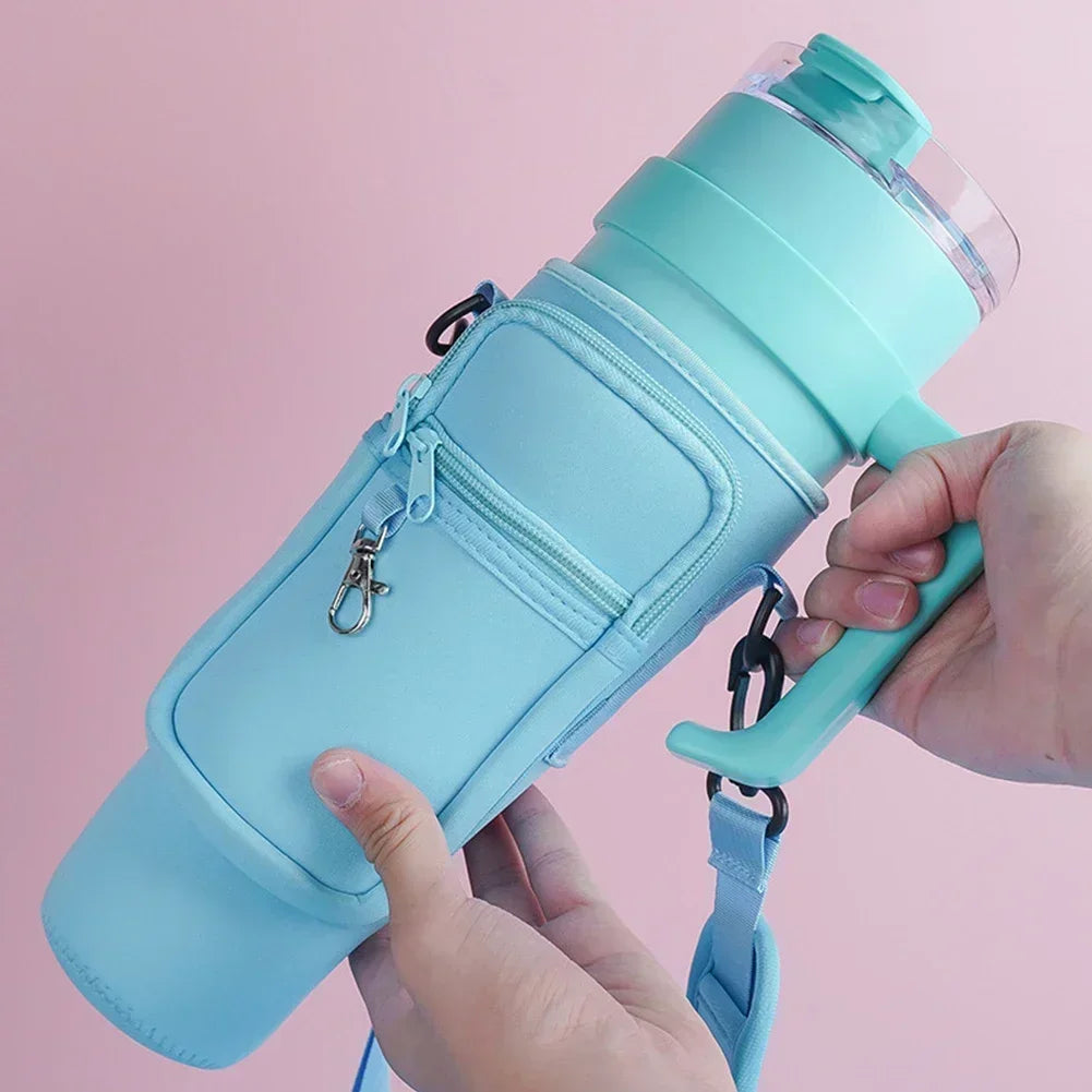 A hand is holding a light blue neoprene water bottle carrier bag with a strap against a pink background.