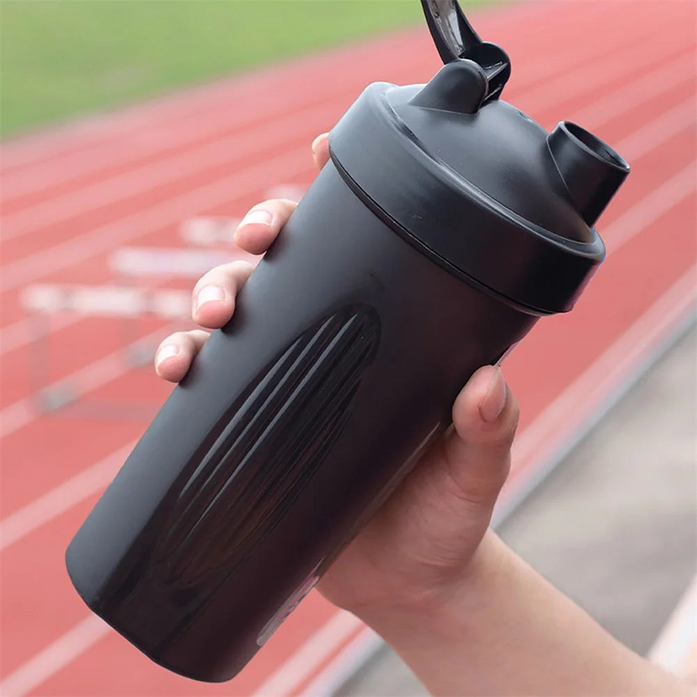 A hand holds a black portable fitness shaker bottle on a red running track surface.