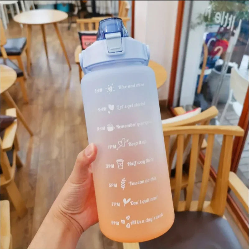 Una mano sostiene una botella deportiva transparente rosa con tapa y pajita azul contra un fondo interior desenfocado.