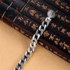 A silver mantra charm bracelet with a textured link chain and a round charm is displayed on a wooden box.