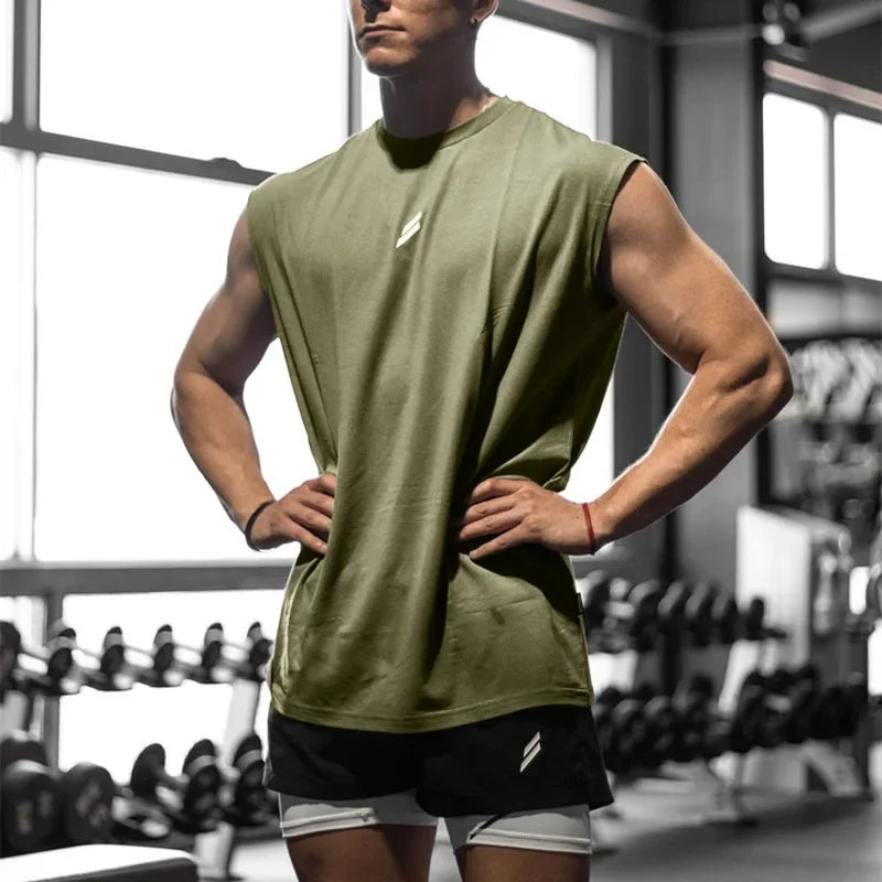 A male model wearing a olive green sleeveless fitness tank top and black shorts in a gym setting.