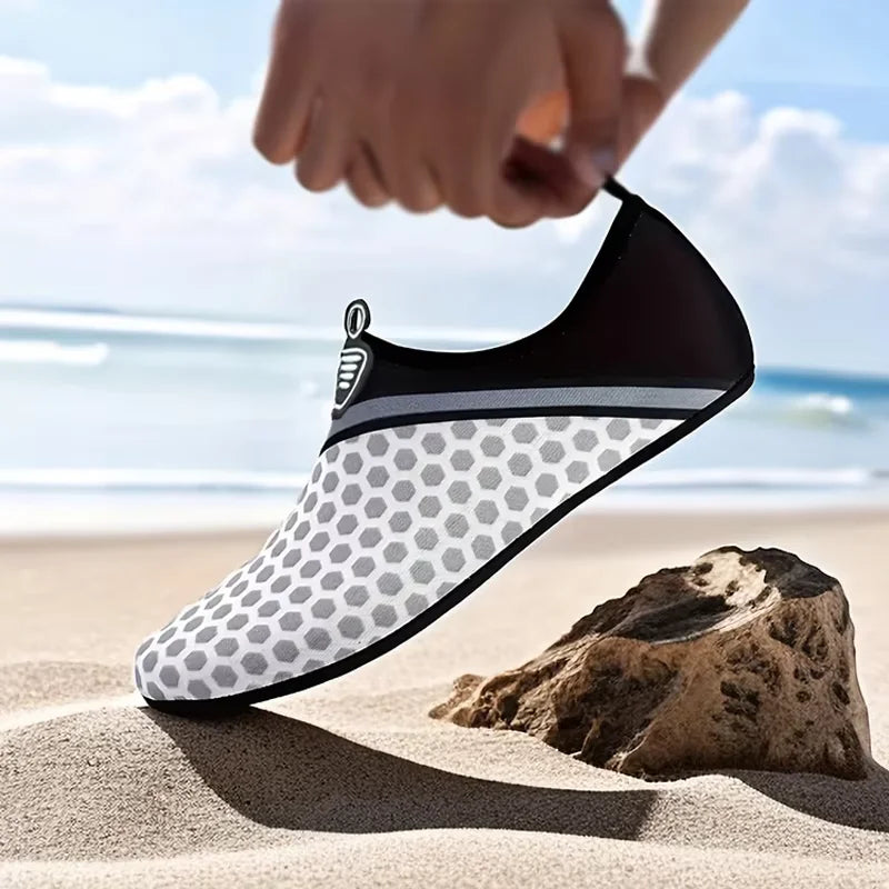 A hand is placing a black and gray waterproof aqua sock on a sandy beach next to a rock.